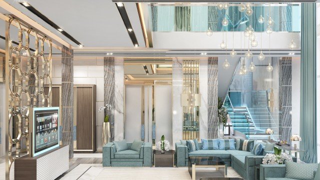 This picture shows a large, ornately decorated living room. The room has a high ceiling with intricate detailing and is painted a warm grey color. The walls are adorned with gold accents, while the floor is covered by a large rug featuring an intricate oriental design. Several luxurious furniture pieces can be seen in the room, including two armchairs upholstered in a vivid blue fabric, and a large sofa framed by glass tables. An elegant chandelier hangs from above, providing an elegant light for the space.