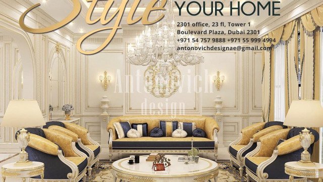 This picture shows a luxurious bedroom designed by Antonovich Design. The room features a large king-sized bed with a tufted velvet headboard and diamond pattern detailing. There is an ornate chandelier directly above the bed, elegant nightstands on either side, and a beautiful round mirror framed in gold leaf. The walls are painted a deep navy blue hue, and a plush white area rug extends from the bed to the seating area in front of the fireplace.