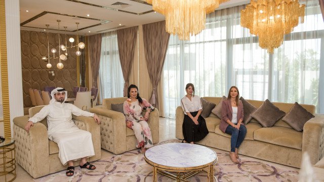 Modern living room furnished with a sectional sofa, armchair, and center table. Several decorative accessories are present, such as wall-mounted art, carpets, and potted plants.