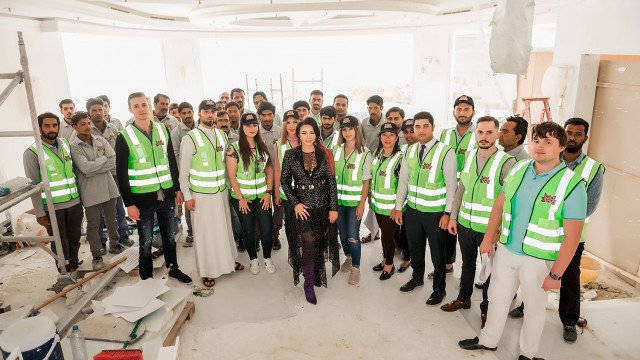In this picture, we can see a luxurious and modern home interior design. The walls are painted in a pale pale pink color, with a white fireplace, and white mantle piece supporting an ornate gold mirror. There is a cream colored patterned rug on the floor, with a black sofa and two matching armchairs, along with a glass topped coffee table. The sofa and chairs are complemented by two sleek modern side tables and side lamps with glass shades. On the wall opposite the fireplace, a large flat-screen television is featured.