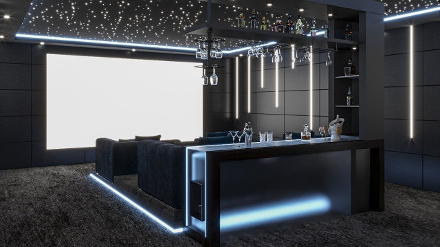 This picture is showing a modern living room with a sectional couch, armchair, and coffee table. The walls are white and the flooring is a combination of wood and patterned tiles. The sectional couch is grey and has a matching ottoman, while the armchair is a deep blue. On the coffee table are some interesting decorative elements, including a colorful vase, a plant, and a bowl filled with pebbles. The lighting comes from a large chandelier hanging above the sectional and two wall sconces.