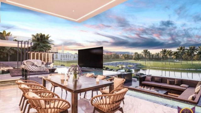 The picture shows a modern luxury living room with luxurious white furniture, plush rugs, and colorful throw pillows. It has a built-in entertainment center with a television and speakers that has shelves for displaying books and other decorations. There is also a large window with a view of the cityscape, as well as a crystal chandelier above the couch and an elegant area rug beneath it.