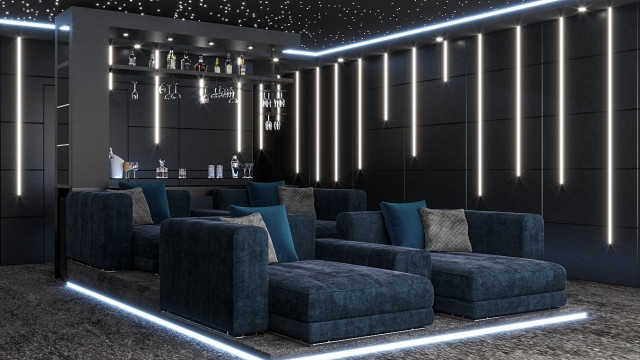 This picture shows a luxurious living room with a semi-circular couch upholstered in soft gray velvet, facing an ornate fireplace. At the center of the space is a large ottoman surrounded by four armchairs with matching upholstery. The walls are decorated with gold accents and there is a large window with sheer curtains and a white marble floor to complete the opulent look.