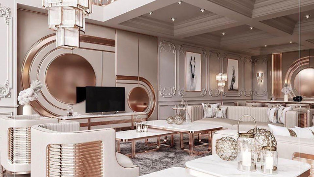 This is a picture of a modern home interior design, featuring a bright, open living room with a high vaulted ceiling, large windows, and a stylish white fireplace. The furniture is modern and minimalistic with two large grey sofas, two white accent chairs, and a low, rectangular coffee table. In the foreground, there is a modern light wood dining table with two mid-century modern dining chairs. In the background, shelves with lights and decorative objects line the walls.