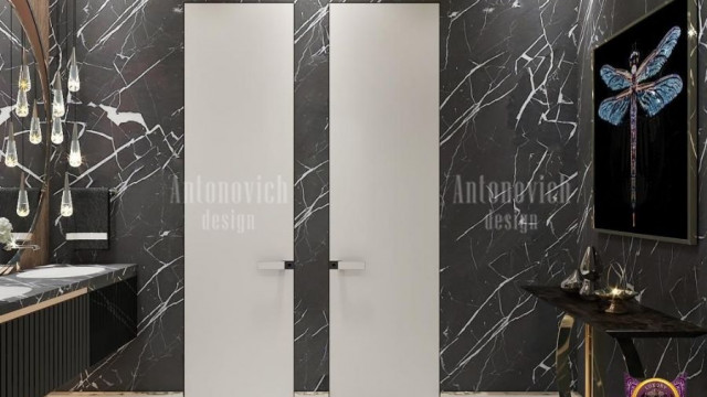 This image shows a luxurious and modern bedroom with grey velvet furniture and a mirrored accent wall. The bed has a light grey velvet headboard, adorned with a large bedspread and multiple round pillows in shades of grey, beige, and white. In front of the bed, there is a wooden nightstand and a light grey velvet armchair with a tufted back. The focal point of the room is the mirrored wall behind the bed, which reflects the entire room, giving it a greater sense of depth and spaciousness.