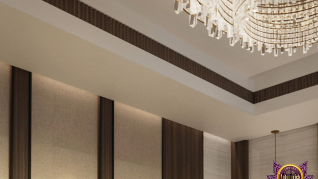 This picture shows a modern interior living room design with sleek and minimalistic furniture, incorporating an off-white sofa, a glass coffee table, and two gray velvet armchairs. It also features a large television mounted to the wall in a black wooden frame, a round work desk, and two decorative plant pots placed on either side of the room. The white walls provide a crisp, bright backdrop for the wood-colored floor and the pieces of furniture.