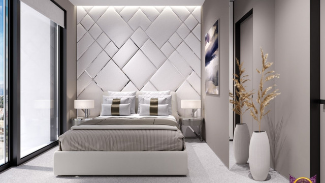 This picture is of a luxurious and modern living room designed by Antonovich Design. In the foreground, there is a circular cream-colored sofa in front of a white coffee table. Behind it is an ornate gold-painted bookcase on either side of the doorway. On the left wall is an ornate mirror flanked by two framed paintings. The walls and floors are a light beige color, with a large area rug in the center of the room. There are also several tall potted plants scattered throughout the space, adding a hint of greenery to the otherwise neutral palette.