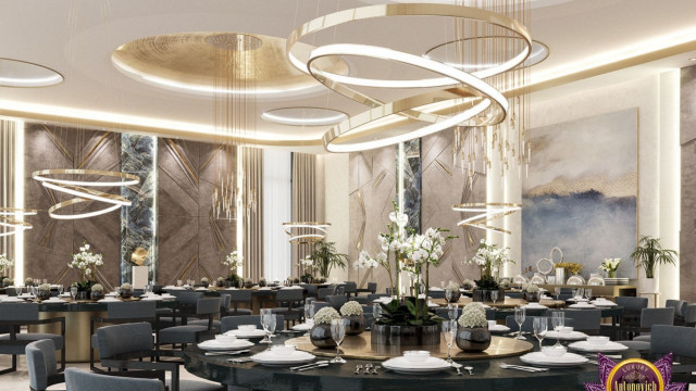 This picture shows a luxurious living room designed by Antonovich Design. It is decorated with modern furniture, gold accents, and crystal chandeliers. The walls are painted in a light grey color, and the floor is covered with a cream-colored rug. There is an ornate fireplace on one wall, and the opposite wall features a large, abstract art piece that adds a touch of sophistication.