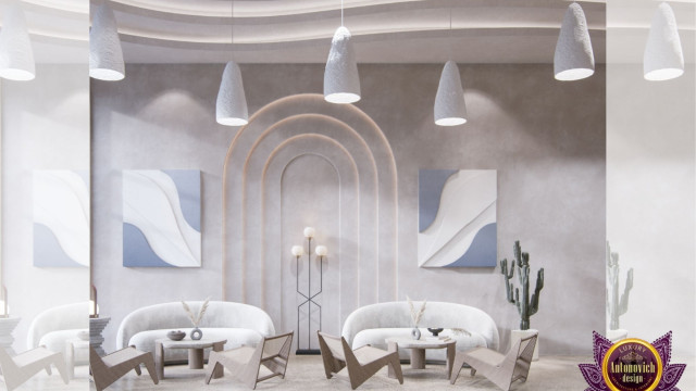 This picture shows a luxurious interior design featuring an open-concept living and dining space. The main focal point appears to be the large white custom-built media wall in the center of the room that houses a state-of-the-art home entertainment system. The furniture is modern and plush, with a velvet maroon sofa and accompanying leather armchairs. The overall look is one of opulence and sophistication, beautifully finished off with hints of gold accenting the ceiling and wall fixtures.