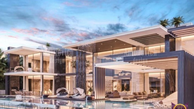 This picture appears to show a contemporary living room with light wood floors and a white ceiling. The walls are painted a soft grey, and the main focus of the room is a white L-shaped sectional sofa with accent pillows in shades of blue and grey. On either side of the sofa are modern white coffee tables with tall metal legs. On one side there is a decorative hearth with white and grey stones and a large metal candle holder. There is also a black media console with two large speakers and a wall-mounted television. The overall design of the room is very modern and