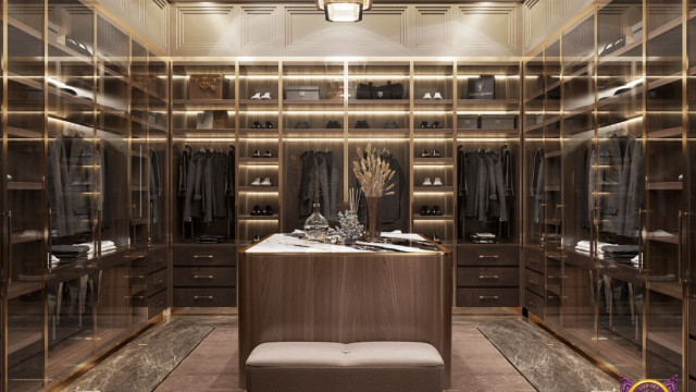 This picture shows a modern bedroom with sophisticated design elements. There is a white bed with tall grey and black headboard, and a white and grey linens. On the sides, there are two nightstands with table lamps and accessories. The walls are painted in a light grey color and on one wall there is a marble accent with a large abstract painting. Above the bed, there is an ornate mirror framed with a black and silver leaf pattern. The floor is laid with a grey carpet and there is a dark rattan armchair in the corner of the room.