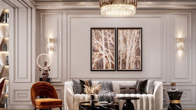 This picture depicts a modern apartment living room, with a sleek and minimalistic design. The room has white walls and furniture, contrasted by elements of dark wood, grey upholstery, and a few touches of greenery. There is an armchair in the corner and a sofa facing a large flat screen television. In the center of the room is a low coffee table where several stylish decor items have been artfully arranged.