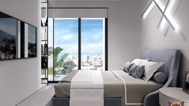 This image is of a modern living room designed by Antonovich Design. The room features a light grey sectional sofa paired with two turquoise accent chairs. The walls are painted a soft white shade, and the ceiling has recessed lighting. A black marble fireplace with a built-in media console is a focal point in the room, and the floor is covered with a light grey carpet. There is a large glass window looking out over a pool and a garden.