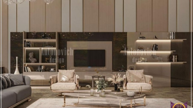 This picture shows a modern and luxurious living room design. It includes a white L-shaped sofa with gold accents, a glass top coffee table, a plush area rug, and a large flat-screen television mounted on the wall. The walls are painted a light gray color and there are several large windows for natural light. At the center of the room is an elegant crystal chandelier with cascading lights.
