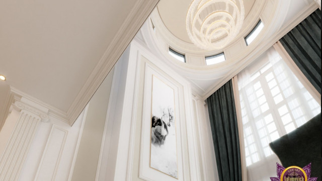 This picture shows a luxurious modern living room with a large beige sectional sofa, an ornate gold coffee table, and a glass top end table. The walls are painted a light grey color and adorned with fine art pieces. There is a unique ceiling light fixture and several small accent lamps. In the background there is a sliding glass door leading out to a balcony with a large brown wicker patio set.