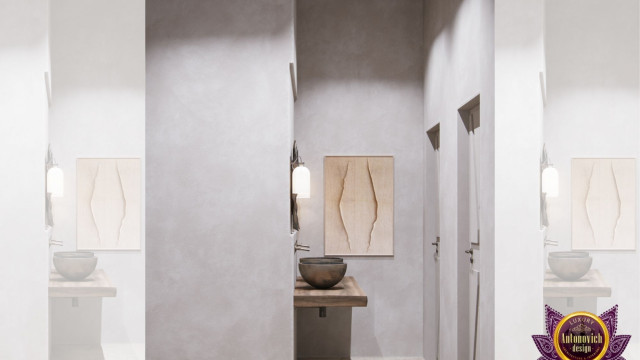 This picture shows a modern and luxurious living room designed by Antonovich Design. It features a light gray sectional sofa with coordinating accent pillows, a small glass top coffee table, and a modern fireplace on the wall. The walls are painted in a warm light gray color, and the ceiling is adorned with beautiful white crown molding. The floor is covered in a large, luxurious white rug, and the space is illuminated with a few strategically placed recessed lights.