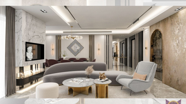 This picture shows a luxurious sitting room in a modern home. The room features a large, curved white sofa along with a round white ottoman in the center of the room. The walls are painted a light shade of blue and are adorned with multiple pieces of art. A striking crystal chandelier hangs from the ceiling and provides light to the entire room. There is also a large fireplace on the wall across from the sofa and several potted plants around the room.