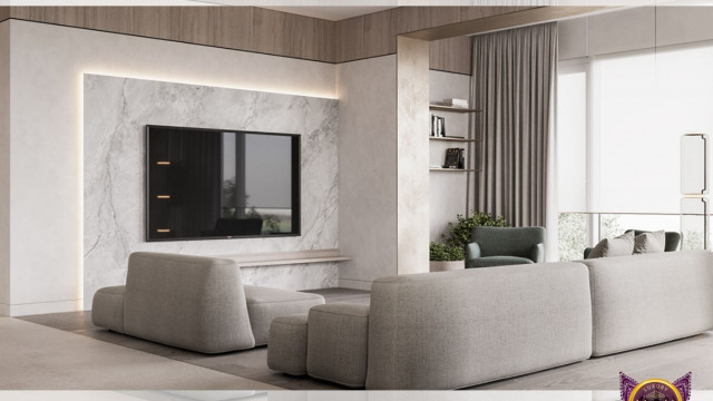 The picture is of a modern living room. It features a white, leather modular sofa arranged in an 'L' shape with a glass-top coffee table in front of it at the corner. There is an abstract painting on the wall, and a white and blue geometric patterned rug covers the floor. The room is lit by two recessed overhead lights and two adjustable wall sconces, and there is a window to the left that provides natural light.