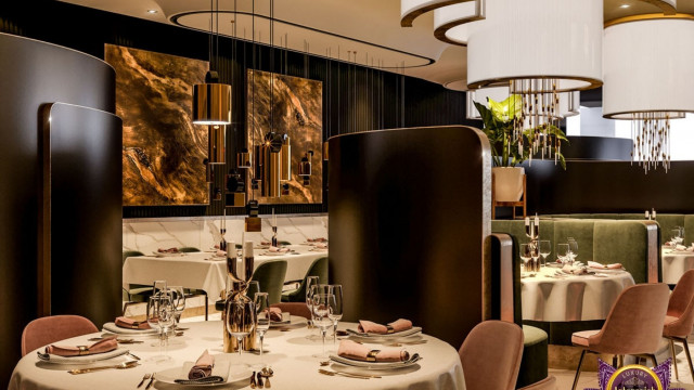 This picture shows two elegant and modern chairs in a luxurious living room. The chairs are cream in color, with tufted detailing along the back. They have slim gold legs and armrests and are placed near a large glass table. The backdrop consists of grey curtains, dark stained wood walls, and a large mirror framed in gold metal.