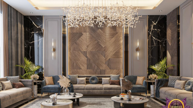 This picture appears to show a modern luxury bedroom design. It features a light gray, tufted headboard and footboard against a white wall decorated with a large silver-framed abstract painting. The bed is topped with two gray pillows, a white duvet and a pink throw blanket. On the left side of the bed is a small white side table with a silver lamp, a gold vase filled with pink roses, and a white decorative box. The floor is covered in a light gray carpet with a pink and beige rug at the foot of the bed.