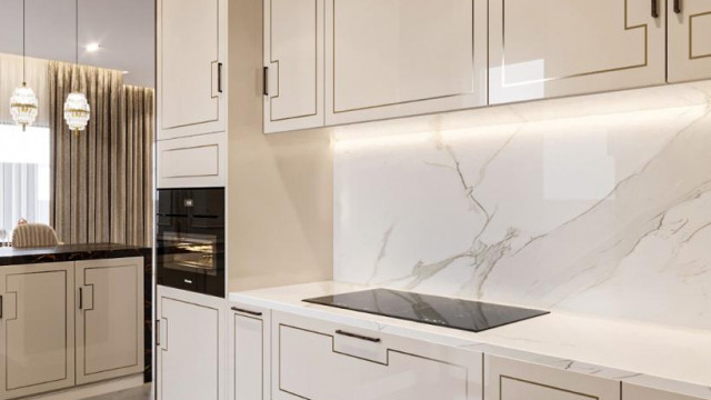 This picture shows a luxurious living room with a large sectional upholstered in white velvet. The walls are light grey in color and feature several paintings and photographs. There are two small mirrored accent tables near the couch, and a large glass coffee table in the center. To the left is a large window with golden curtains, overlooking a stunning view of the city skyline. The room is accented with luxurious touches such as a crystal chandelier and golden lamps.