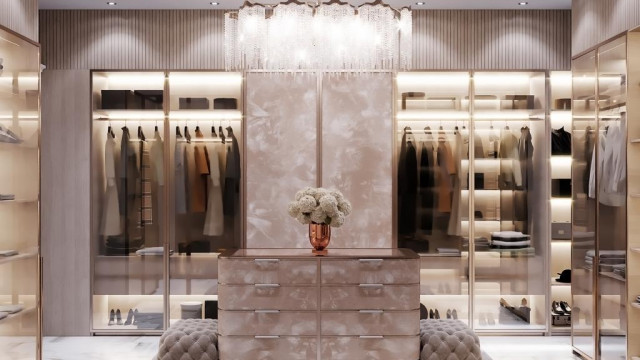 This picture shows a luxurious and modern living room interior. The room is decorated in shades of grey and white, with furniture upholstered in velvet. There is an ornately carved fireplace mantel and a large floor-to-ceiling window that allows plenty of natural light to enter the room. The space is also furnished with a crystal chandelier, a plush sofa, and two armchairs.