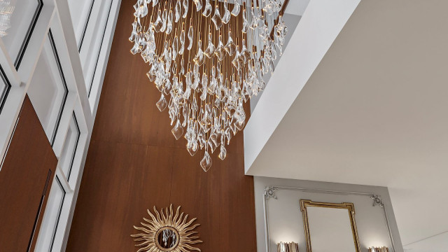 This picture shows a luxurious, traditional living room designed by Antonovich Design. It features golden chandeliers, a white sofa set with patterned cushions, a coffee table with several decorative items on top, and a rug with a golden border in the center. The walls are decorated with ornate furniture pieces and artwork, and the floor is covered in an elegant tiled pattern.
