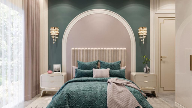 This picture shows a modern living room with a multi-faceted design. The room features dark hardwood floors, an eye-catching white sectional sofa, and a patterned rug. The walls are a matte black color and there is a brown leather ottoman in the center of the room. An off-white armchair sits against the wall and a large mirror hangs above the couch. A set of sliding glass doors lead to a balcony, and there is a recessed light in the ceiling.