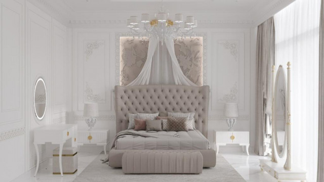 The picture shows an elegant bedroom with a four-poster bed in the center. The walls are painted a pale pink and have ornately detailed wainscoting. The large windows are framed by white curtains and allow natural light to flood the room. The floor is covered in a rich patterned rug, while the bed is draped in a cream duvet and adorned with black and white pillows. A crystal chandelier hangs from the high ceiling, adding a touch of glamour.