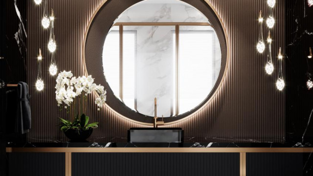 This picture shows a luxurious gold-accented bathroom. There is a free-standing bathtub in the center with a dark brown wooden base and a beige tiled exterior. On either side of the tub are two large, arched windows with white shutters that let in plenty of natural light. The walls of the room are decorated with an ornate white wallpaper and a large gold-framed mirror hangs in the center. There is a modern sink beneath the mirror with two tall gold taps and a marble countertop. Finally, the flooring is a light grey marble with