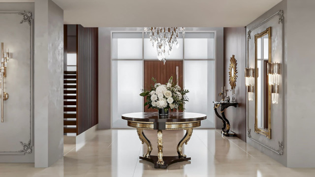 This image shows a luxury master bedroom designed by Antonovich Design. The room features an off-white and gold color palette, with ornate architectural detailing and furniture throughout. It includes a large double bed with a canopy, a chaise lounge chair, and a matching dresser. There are also several tall pillars along one wall, and a decorative fireplace near the center of the room. Large floor-to-ceiling windows let in plenty of natural light, and the room is filled with lush greenery and warm lighting.