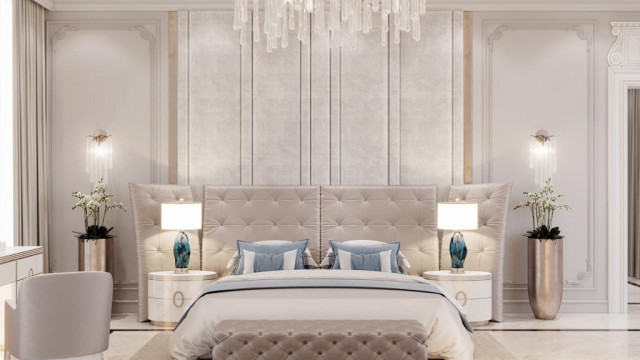 This picture is of a luxurious living room designed by Antonovich Design. The room features high ceilings with multiple elegant glass chandeliers, an ornately decorated fireplace, and a large area rug that gives the space a warm, inviting atmosphere. The walls are decorated with large canvas paintings and detailed moldings, and the furniture includes two sofas, an armchair, and two round tables. Numerous side tables and lamps provide additional lighting and decor.