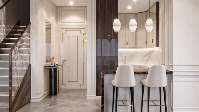 This picture shows a luxurious living room with modern furniture, warm colors, and elegant touches. The room features a beige sofa and two armchairs upholstered in a geometric pattern, along with a deep red accent chair. The walls are decorated with framed artwork, while a stately fireplace makes a focal point. A vintage lamp sits atop an end table, and various knick-knacks decorate the shelves on either side. The floor is covered in hardwood, and a large area rug adds warmth to the inviting space.