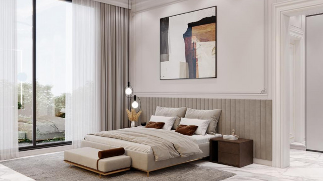 This picture shows a luxurious bedroom design with a large, gray upholstered headboard with tufting and gold accents. The bed is surrounded by floor to ceiling windows with sheer curtains for natural light. There is an area rug on the hardwood floor and a pair of armchairs beside the bed. There are also gold accents in the art, lamps, and furniture, creating a warm and inviting atmosphere.