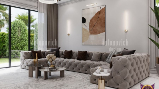 This picture shows a luxurious living room in an elegant style, featuring two white and gold sofas with matching armchairs and a glass coffee table. The walls are adorned with ornate patterned wallpaper in muted colors, and a grand carved wooden fireplace adds warmth to the space. Large windows let in plenty of natural light and lead out onto a balcony space with a view. The overall look is sophisticated and inviting.