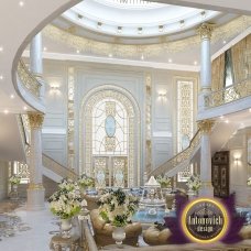 This picture shows a large, white building with four rectangular windows and an arched entrance. There is a grey stone staircase leading up to the entrance, and a metal railing around it. In the background, there are trees and green shrubs.